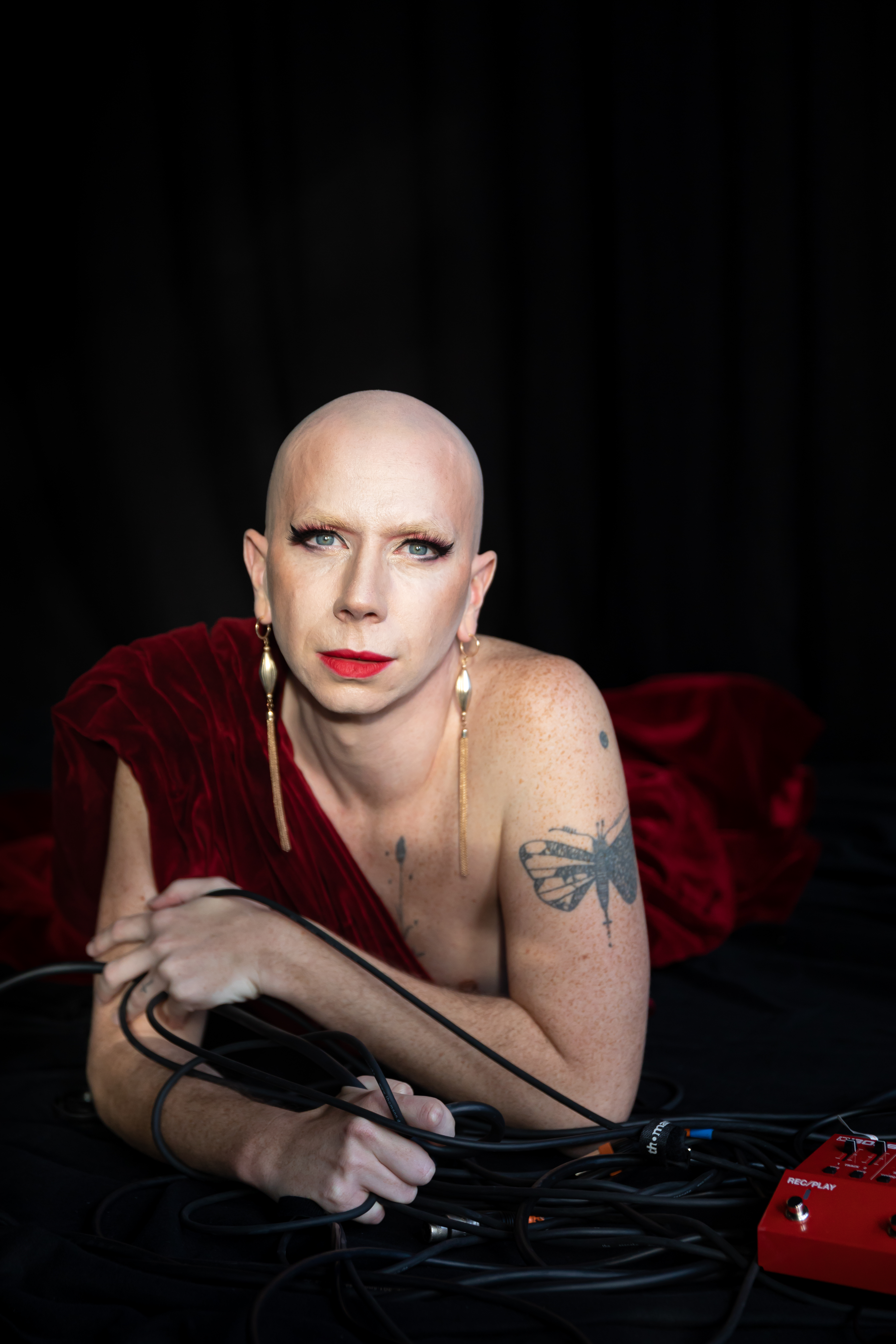 Olympia Bukkakis Olympia Bukkakis trägt knallroten Lippenstift, liegt auf dem Boden in einer samtroten Robe vor schwarzem Hintergrund.