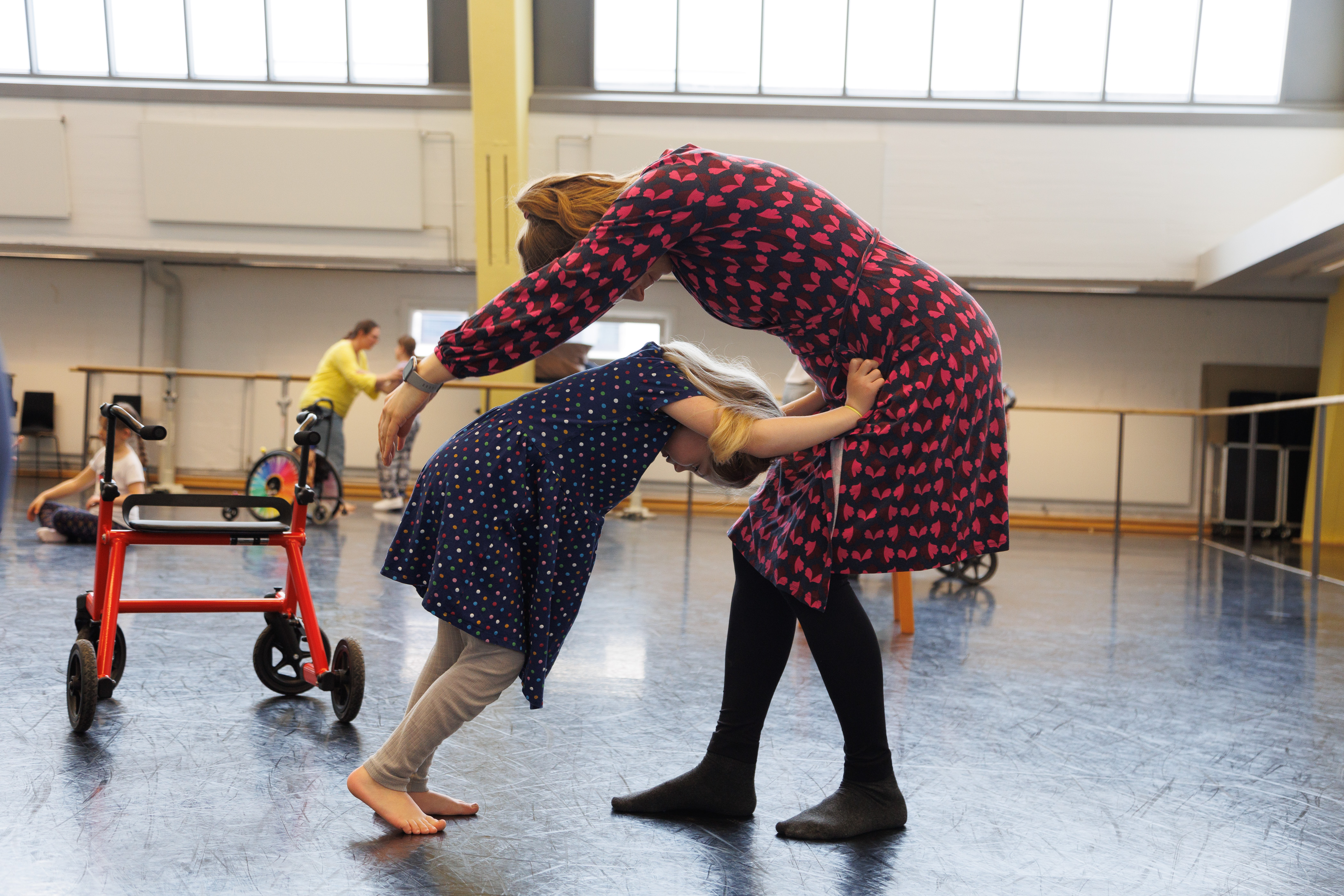Kind und Mutter stehend im Ballettsaal, Kind stützt sich gebeugt auf den Oberschenkeln der Mutter ab, Mutter umschließt Kind hinter dem Rücken
