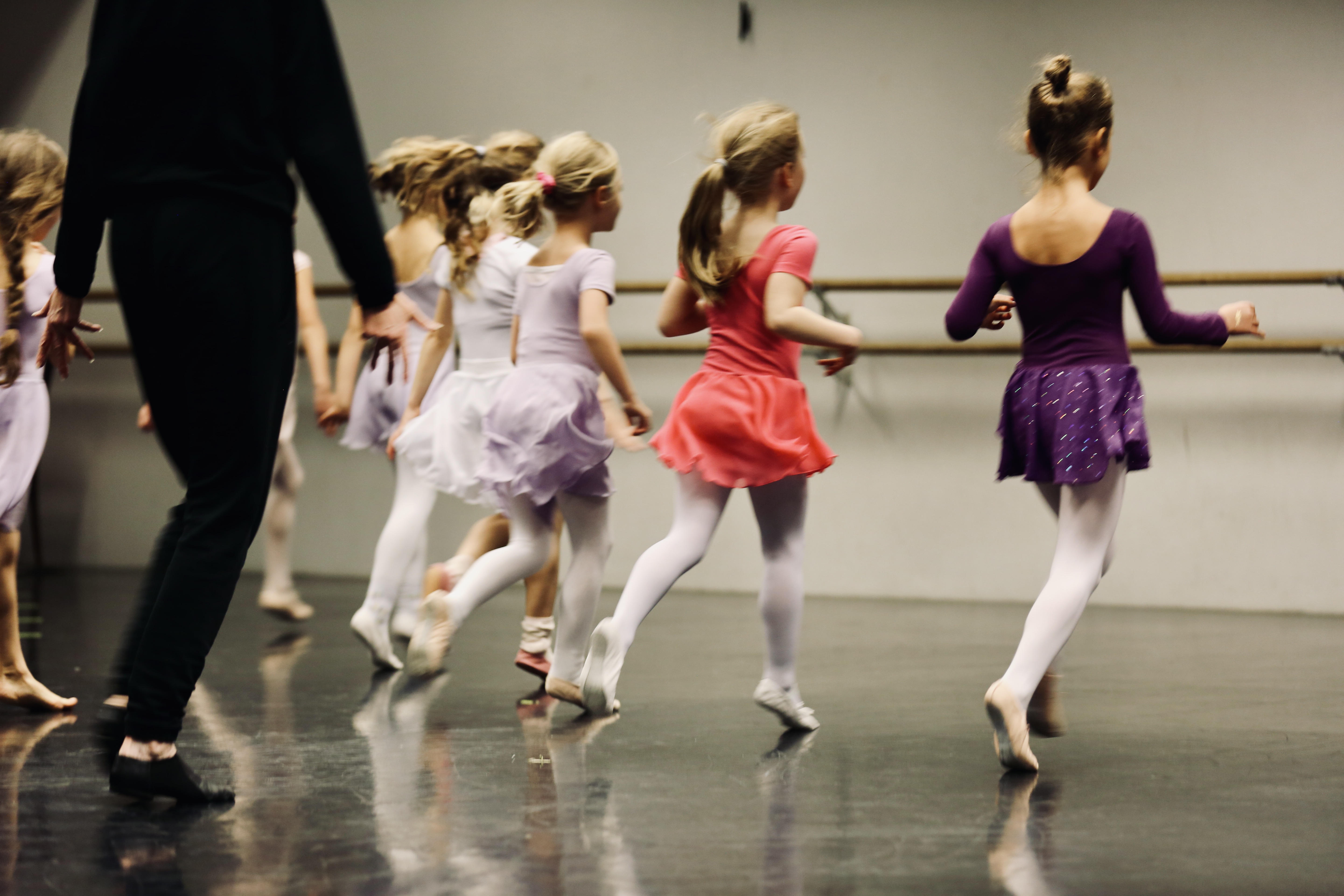 Vier Kinder in halbnaher Aufnahme, die mit dem Rücken zur Kamera in einem Ballettsaal Richtung Ballettstange rennen. Hinter ihnen ist der Körper der Ballettlehrerin zu sehen.