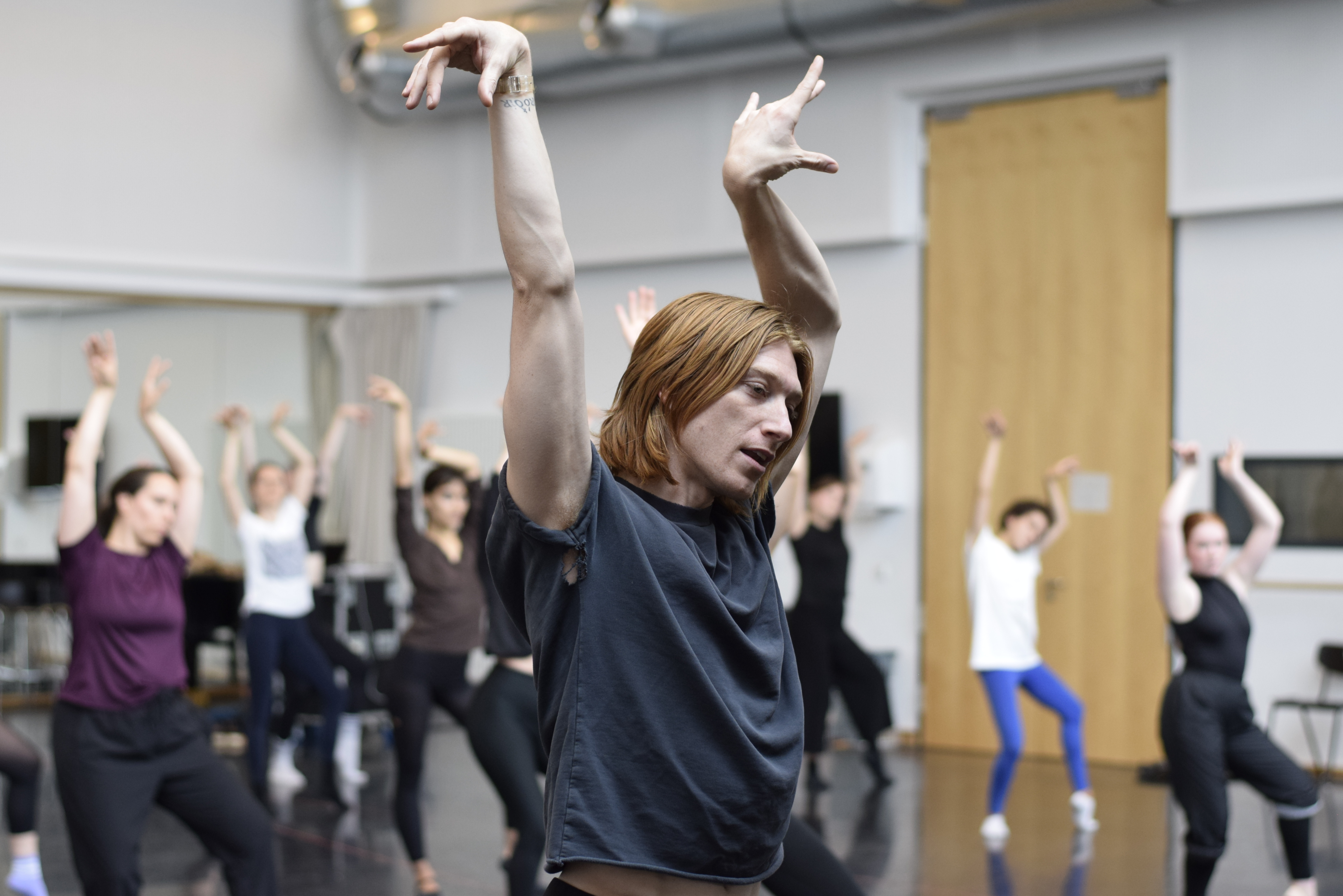 Im Vordergrund halbnah ein Tänzer in Pose mit erhobenen Händen, Kopf und blick seitlich nach unten geneigt. Im Hintergrund unscharf sind weitere Tänzerinnen zu sehen, in ähnlicher Pose.