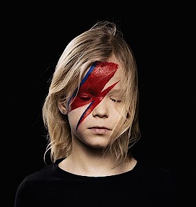 Headshot eines blonden Mädchens mit geschlossenen Augen vor schwarzem Hintergrund. Auf ihrer rechten Gesichtshälfte ist ein roter Blitz geschminkt.