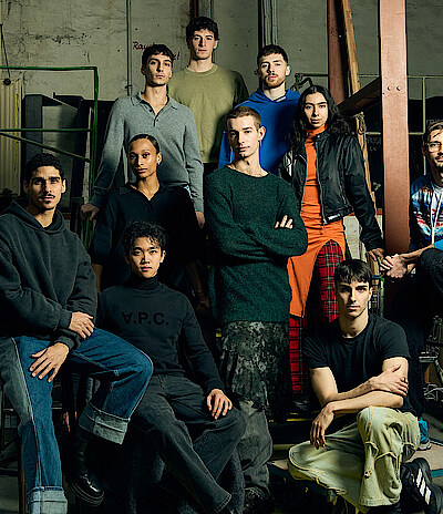 Gruppenfoto der elf Next Generation Choreograph*innen im Lager der Deutschen Oper Berlin, umgeben von Holzrahmen, Kisten und Rollwägen.