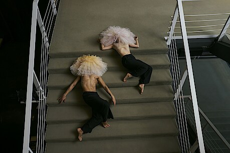 Zwei Tänzer*innen mit Tutu auf dem Kopf liegen auf einer Treppe mit Teppichbelag.