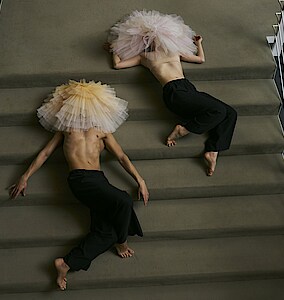 Spielzeit 24/25 Zwei Tänzer*innen mit Tutu auf dem Kopf liegen auf einer Treppe mit Teppichbelag.