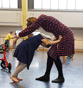 Kind und Mutter stehend im Ballettsaal, Kind stützt sich gebeugt auf den Oberschenkeln der Mutter ab, Mutter umschließt Kind hinter dem Rücken