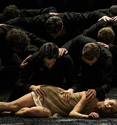 Weronika Frodyma liegt auf dem Boden, Tänzer in schwarzen Kostümen beugen sich über sie.