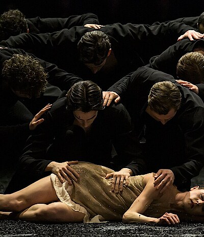 Weronika Frodyma liegt auf dem Boden, Tänzer in schwarzen Kostümen beugen sich über sie.