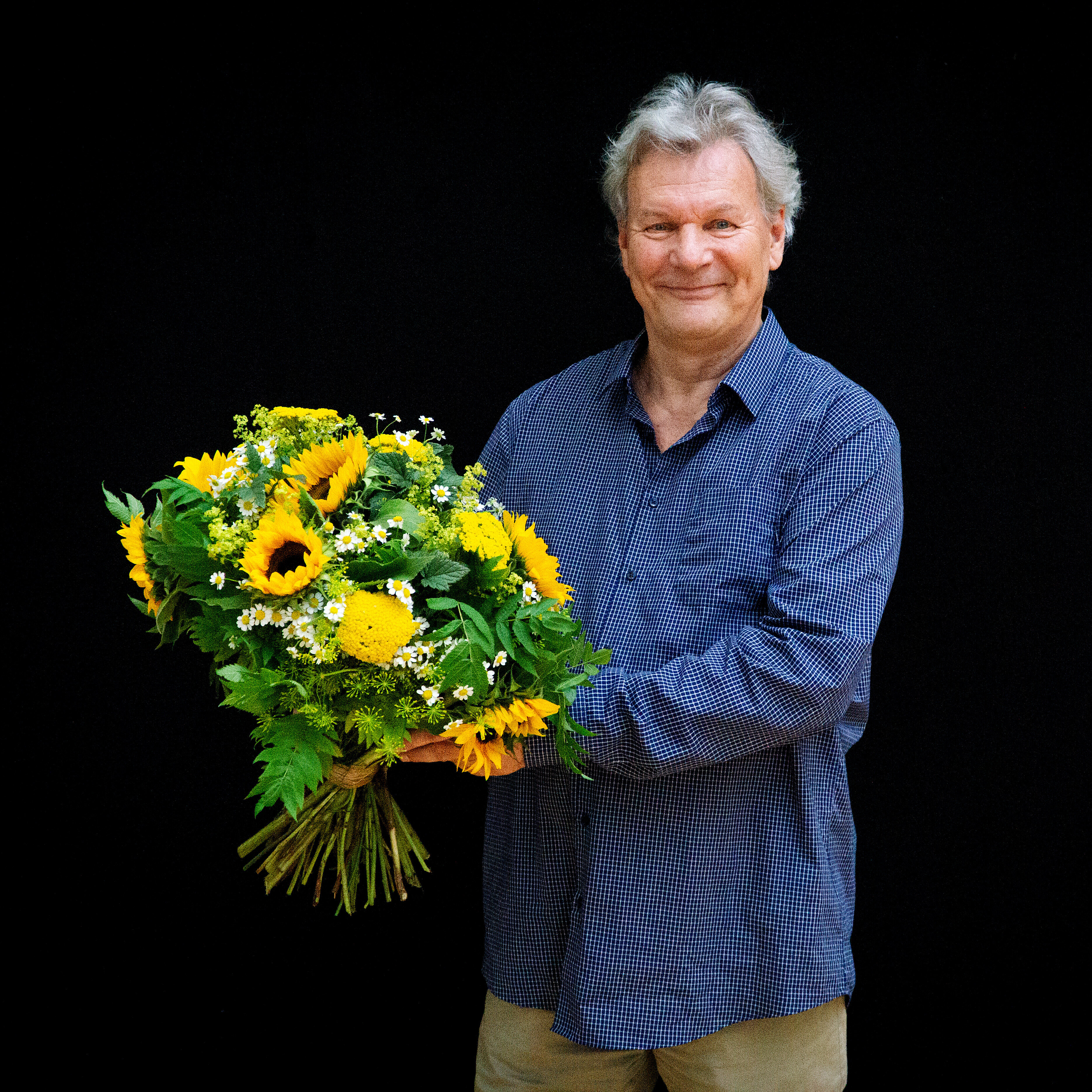 Portrait Peter Hartwig in blaukariertem Hemd vor schwarzem Hintergrund. Er hält vor sich einen Strauß mit Sonnenblumen und lächelt in die Kamera.