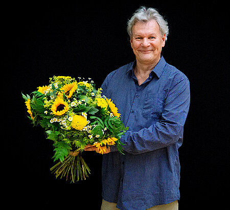 Portrait Peter Hartwig in blaukariertem Hemd vor schwarzem Hintergrund. Er hält vor sich einen Strauß mit Sonnenblumen und lächelt in die Kamera.