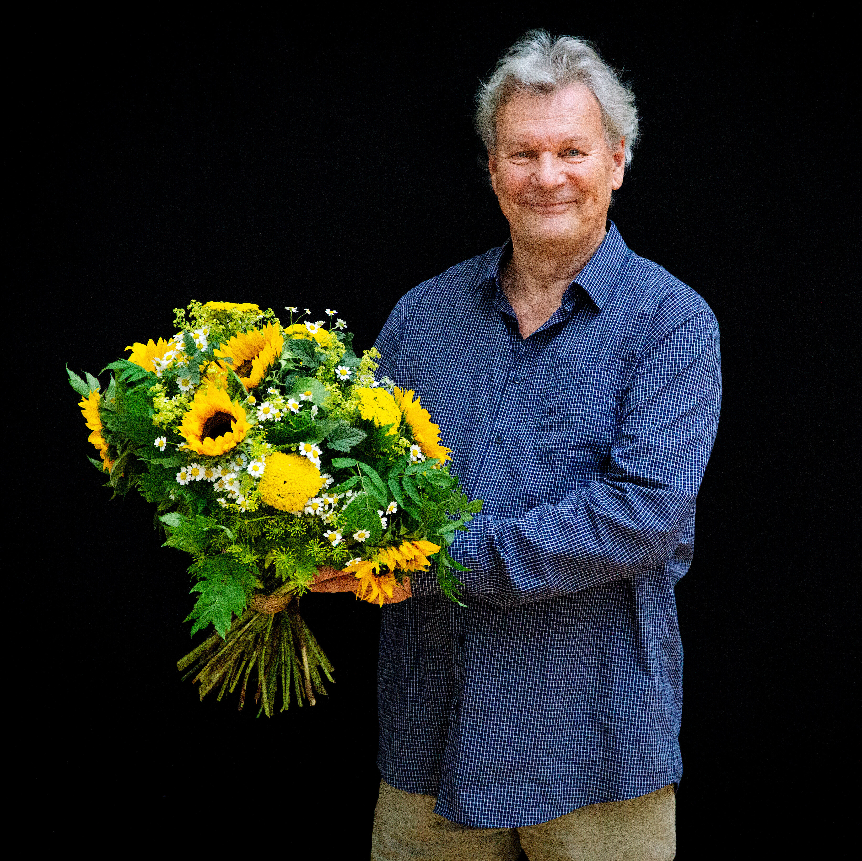 Portrait Peter Hartwig in blaukariertem Hemd vor schwarzem Hintergrund. Er hält vor sich einen Strauß mit Sonnenblumen und lächelt in die Kamera.