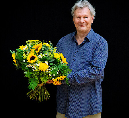 Portrait Peter Hartwig in blaukariertem Hemd vor schwarzem Hintergrund. Er hält vor sich einen Strauß mit Sonnenblumen und lächelt in die Kamera.