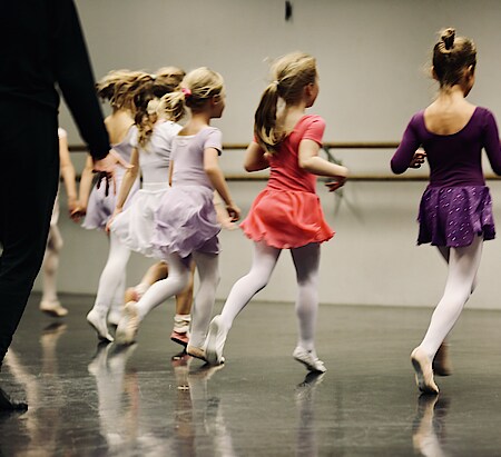 Vier Kinder in halbnaher Aufnahme, die mit dem Rücken zur Kamera in einem Ballettsaal Richtung Ballettstange rennen. Hinter ihnen ist der Körper der Ballettlehrerin zu sehen.