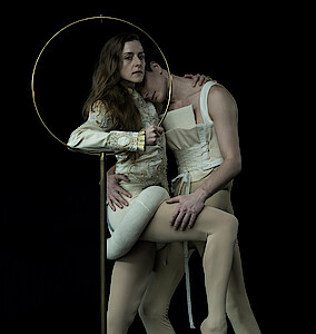 Weronika Frodyma und Jan Casier tragen beige Korsette vor schwarzem Hintergrund und halten sich im Arm. Weronika stützt sich auf ein goldenes Metallgestellt.
