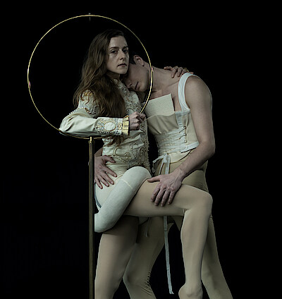 Weronika Frodyma und Jan Casier tragen beige Korsette vor schwarzem Hintergrund und halten sich im Arm. Weronika stützt sich auf ein goldenes Metallgestellt.