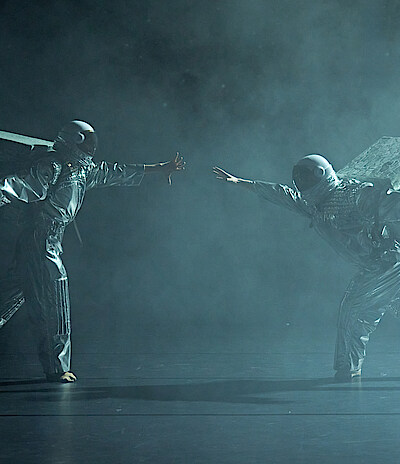 Zwei Astronaut*innen in silbernen Raumanzügen vor schwarzem Hintergrudn bewegen sich durch Bühnennebel mit ausgestreckten Händen aufeinander zu.