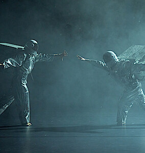 Zwei Astronaut*innen in silbernen Raumanzügen vor schwarzem Hintergrudn bewegen sich durch Bühnennebel mit ausgestreckten Händen aufeinander zu.