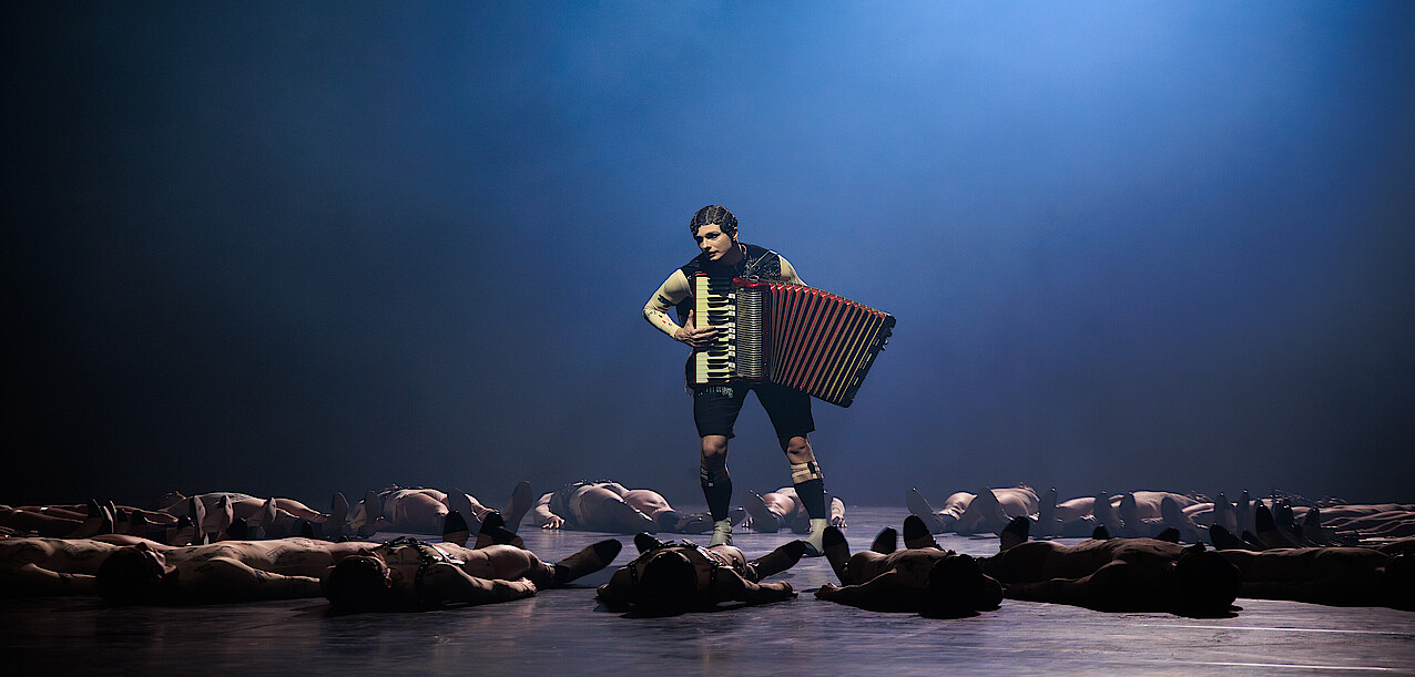 Jan Casier steht in einem vernebelten Lichtkegel. Er trägt eine schwarze Weste und kurze Hose. Er steht gekrümmt und hat ein Akkordeon umgeschnallt. Er blickt verloren ins Weite. Um ihn herum liegen Tänzer*innen im Kreis. 