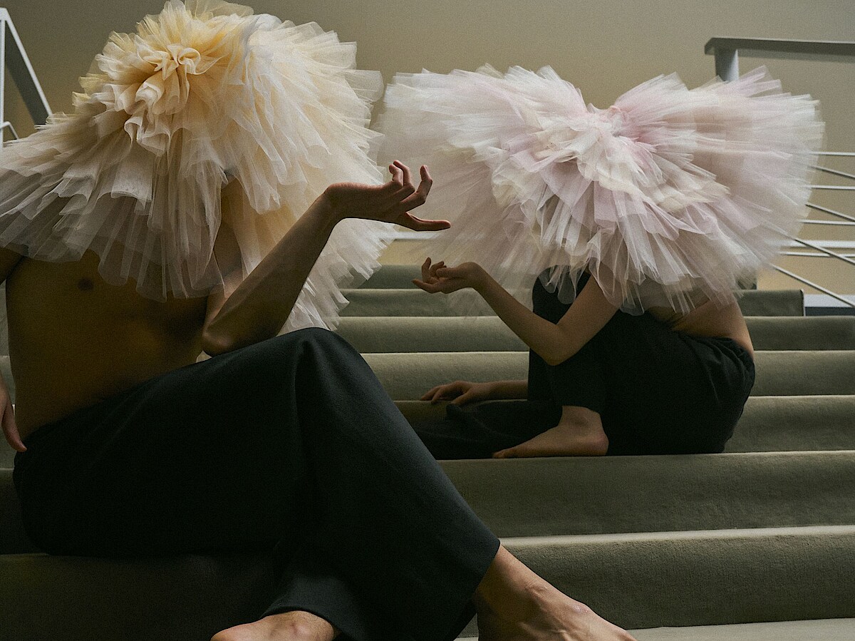 Ballett Gala Zwei Tänzer*innen mit Tutus auf dem Kopf sitzen auf einer Treppe mit grauem Teppichbelag