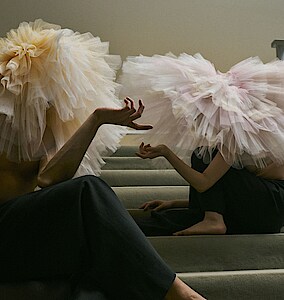 Zwei Tänzer*innen mit Tutus auf dem Kopf sitzen auf einer Treppe mit grauem Teppichbelag