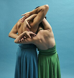 Zwei Tänzer*innen in Röcken stehen mit freien Rücken zur Kamera und haben ihre Arme verschränkt