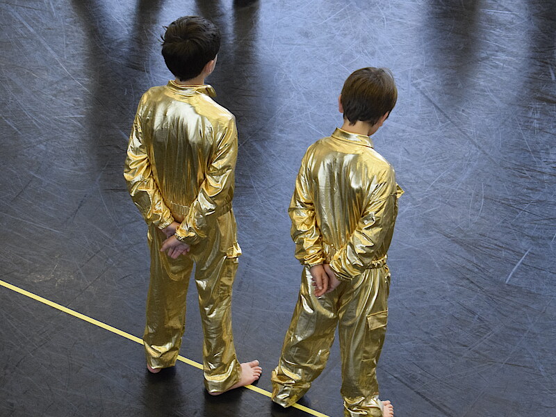 Fotografiert von schräg oben sind zwei Kinder von hinten im Ballettsaal zu sehen. Sie stehen breitbeinig und tragen goldene Ganzkörperanzüge