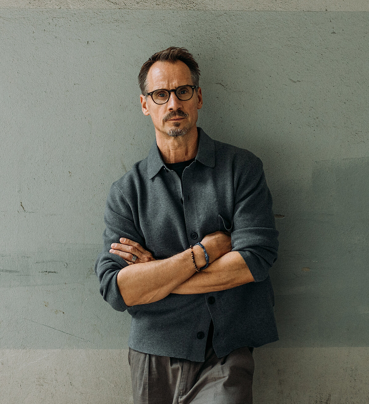 Christian Spuck in grauem Hemd lehnt mit dem Rücken an einer grau verputzten Mauer, die Arme vor der Brust verschränkt.