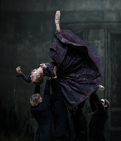 Tänzergruppe in schwarzen Anzügen trägt Weronika Frodyma als Emma Bovary, die ihren rechten Fuß in die Höhe streckt. 