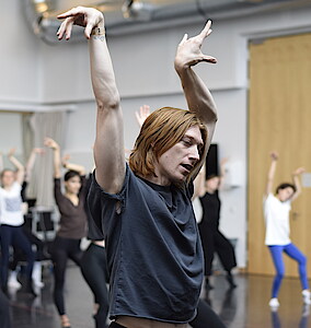 Im Vordergrund halbnah ein Tänzer in Pose mit erhobenen Händen, Kopf und blick seitlich nach unten geneigt. Im Hintergrund unscharf sind weitere Tänzerinnen zu sehen, in ähnlicher Pose.