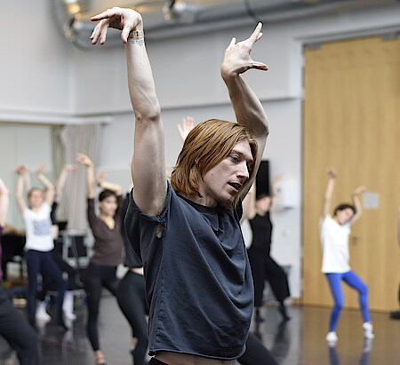 Im Vordergrund halbnah ein Tänzer in Pose mit erhobenen Händen, Kopf und blick seitlich nach unten geneigt. Im Hintergrund unscharf sind weitere Tänzerinnen zu sehen, in ähnlicher Pose.