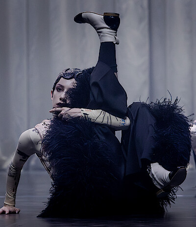 Zwei weiß geschminkte Tänzer*innen tragen schwarze fellige Hosen und einen engen hautfarbenen Body. Sie sitzen auf dem Boden, ein Bein angewinkelt, das andere in die Höhe gestreckt. Im Hintergrund ein grauer Vorhang.