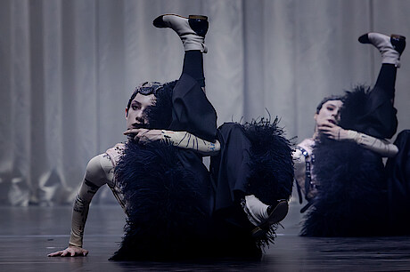 Zwei weiß geschminkte Tänzer*innen tragen schwarze fellige Hosen und einen engen hautfarbenen Body. Sie sitzen auf dem Boden, ein Bein angewinkelt, das andere in die Höhe gestreckt. Im Hintergrund ein grauer Vorhang.