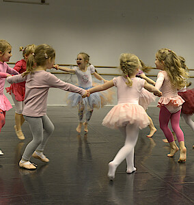 Sieben rosa und pinkt gekleidete Kinder tanzen im Ballettsaal. Sie halten sich an den Händen und bilden einen Kreis.