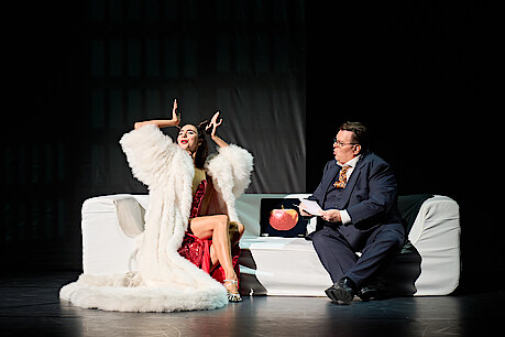 Tänzerin in rotem Abendkleid und weißem Pelzmantel sitzt auf einer weißen Couch und wird von einem Moderator in dunkelblauem Anzug interviewt.