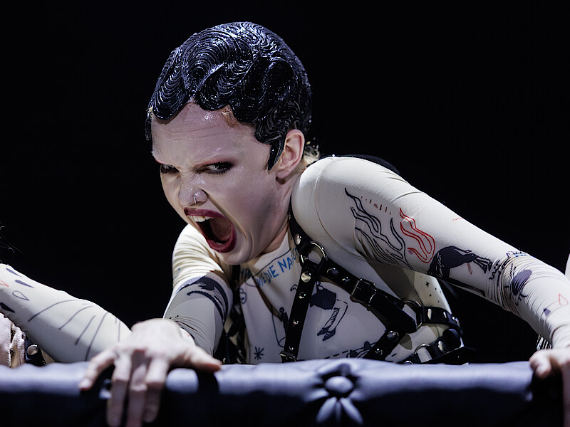 Wunderkammer Großaufaufnahme von Inara Wheeler in weißem Makeup und glänzender Kurzhaarperücke, die sich am schwarz gepolsterten Bühnenrand festhält und grotesk in die Kamera blickt.