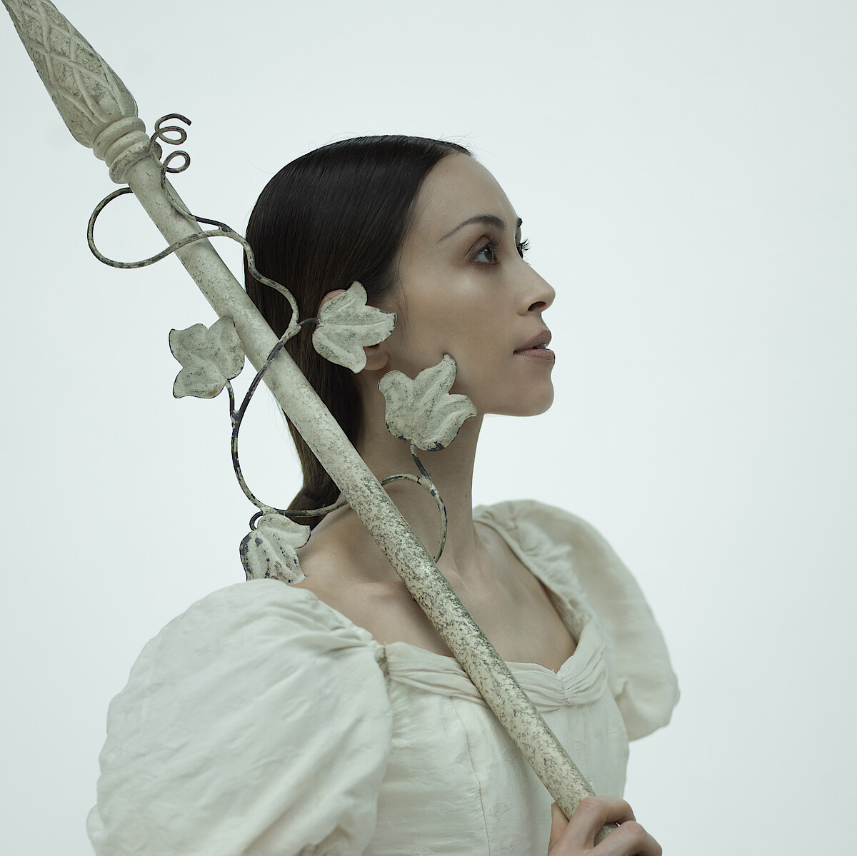 Yoko Callegari trägt ein weißes Kleid und blickt nach rechts oben vor weißem Hintergrund. Über der Schulter trägt sie einen weißen Metallspeer mit Rosen verziert.
